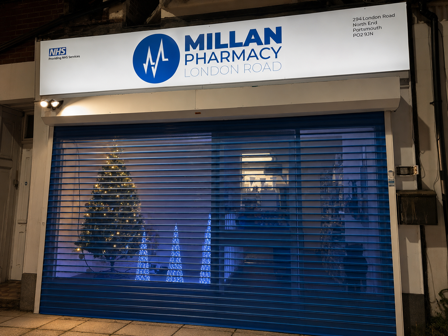 Millan Pharmacy London Road storefront at night with festive window display